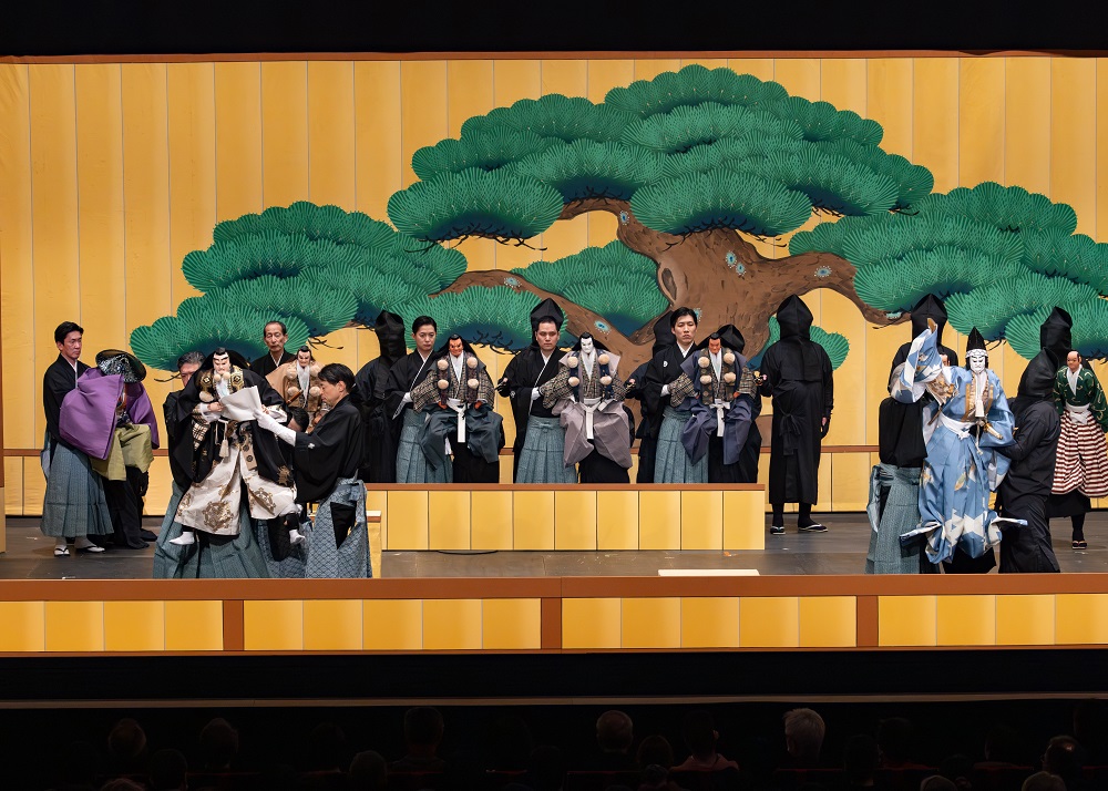 写真：よしつねを後ろに隠し、勧進帳に見立てた白紙の巻物を読む、べんけいと、その様子を見つめる、とがし